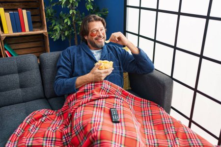 Foto de Hombre de mediana edad con parches debajo de los ojos viendo películas comiendo papas fritas en casa - Imagen libre de derechos