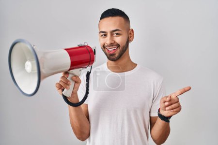 Téléchargez les photos : Jeune homme hispanique criant à travers mégaphone souriant heureux pointant avec la main et le doigt vers le côté - en image libre de droit