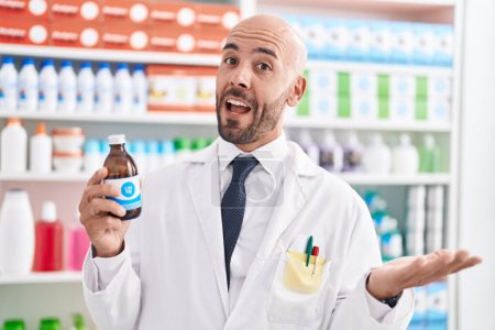 Téléchargez les photos : Middle age bald man working at pharmacy drugstore holding syrup celebrating achievement with happy smile and winner expression with raised hand - en image libre de droit