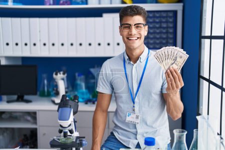 Téléchargez les photos : Young hispanic man working at scientist laboratory holding dollars looking positive and happy standing and smiling with a confident smile showing teeth - en image libre de droit