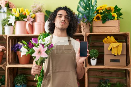 Téléchargez les photos : Homme hispanique avec les cheveux bouclés travaillant à la boutique de fleuriste montrant écran smartphone regardant la caméra souffler un baiser étant belle et sexy. expression d'amour. - en image libre de droit