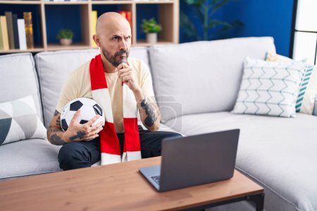 Foto de Hispanic man with tattoos watching football match hooligan holding ball on the laptop serious face thinking about question with hand on chin, thoughtful about confusing idea - Imagen libre de derechos