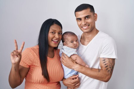 Photo for Young hispanic couple with baby standing together over isolated background smiling looking to the camera showing fingers doing victory sign. number two. - Royalty Free Image