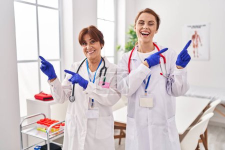Photo for Two women wearing doctor uniform and stethoscope smiling and looking at the camera pointing with two hands and fingers to the side. - Royalty Free Image