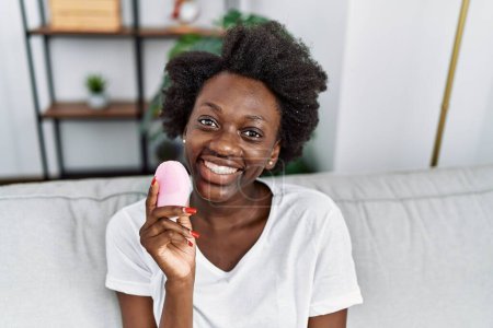 Téléchargez les photos : Jeune femme afro-américaine souriante confiante en utilisant un nettoyant facial électrique à la maison - en image libre de droit