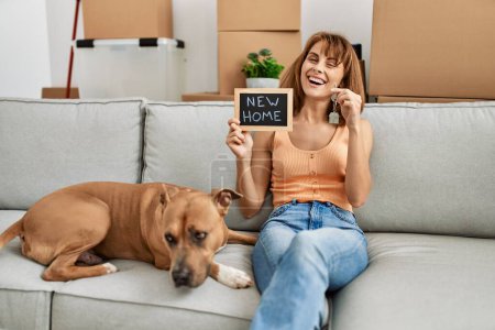 Foto de Joven mujer caucásica sosteniendo la llave y la nueva pizarra casera sentada en el sofá con el perro en casa - Imagen libre de derechos