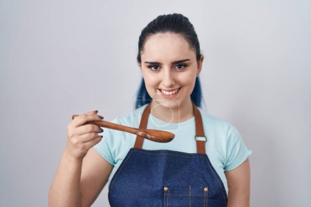 Téléchargez les photos : Young modern girl with blue hair wearing cook apron holding spoon looking positive and happy standing and smiling with a confident smile showing teeth - en image libre de droit