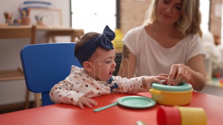 Photo for Woman and toddler learning to eat with plastic dish toy sitting on table at kindergarten - Royalty Free Image