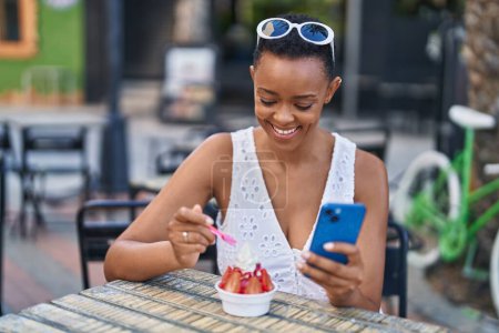 Photo for African american woman eating ice cream using smartphone at coffee shop terrace - Royalty Free Image