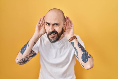 Foto de Young hispanic man with tattoos standing over yellow background trying to hear both hands on ear gesture, curious for gossip. hearing problem, deaf - Imagen libre de derechos