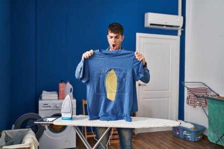 Foto de Young hispanic man ironing holding burned iron shirt at laundry room in shock face, looking skeptical and sarcastic, surprised with open mouth - Imagen libre de derechos