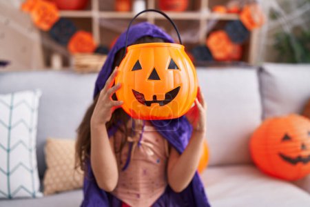 Foto de Adorable hispanic girl wearing halloween costume holding pumpkin basket over face at home - Imagen libre de derechos