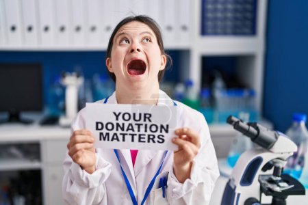 Foto de Woman with down syndrome working at scientist laboratory holding your donation matters banner angry and mad screaming frustrated and furious, shouting with anger looking up. - Imagen libre de derechos