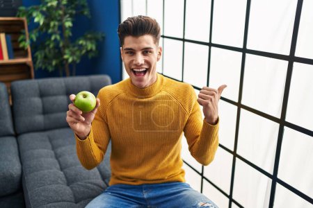 Photo for Young hispanic man holding green apple pointing thumb up to the side smiling happy with open mouth - Royalty Free Image