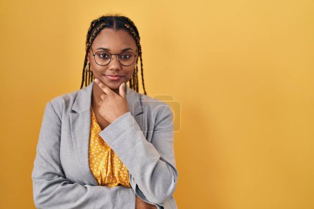 Photo for African american woman with braids standing over yellow background looking confident at the camera with smile with crossed arms and hand raised on chin. thinking positive. - Royalty Free Image