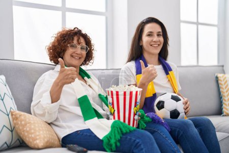 Téléchargez les photos : Mère et fille hispaniques regardant le football soutenir l'équipe souriant heureux et positif, pouce levé faire excellent et signe d'approbation - en image libre de droit