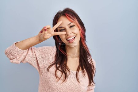 Foto de Young caucasian woman wearing pink sweater over isolated background doing peace symbol with fingers over face, smiling cheerful showing victory - Imagen libre de derechos