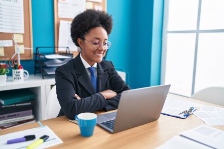 Téléchargez les photos : Femme afro-américaine travailleur d'affaires en utilisant un ordinateur portable assis avec les bras croisés geste au bureau - en image libre de droit