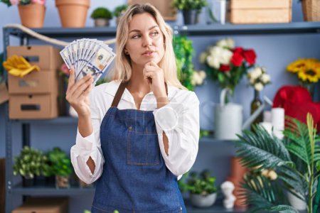 Photo for Young blonde woman working at florist shop holding dollars serious face thinking about question with hand on chin, thoughtful about confusing idea - Royalty Free Image