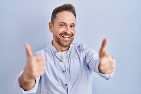 Photo for Middle age caucasian man standing over blue background approving doing positive gesture with hand, thumbs up smiling and happy for success. winner gesture. - Royalty Free Image