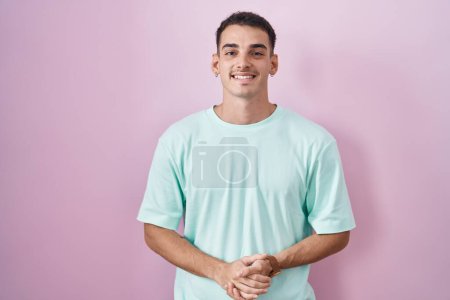 Foto de Hombre hispano guapo de pie sobre fondo rosa con las manos juntas y los dedos cruzados sonriendo relajado y alegre. éxito y optimismo - Imagen libre de derechos
