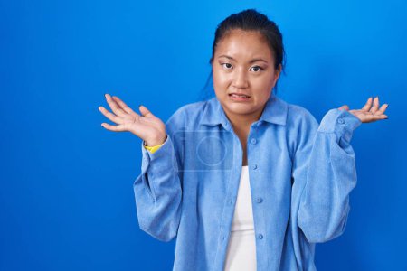 Photo for Asian young woman standing over blue background clueless and confused expression with arms and hands raised. doubt concept. - Royalty Free Image