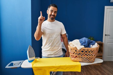 Foto de Young hispanic man with beard ironing clothes at home smiling with an idea or question pointing finger up with happy face, number one - Imagen libre de derechos