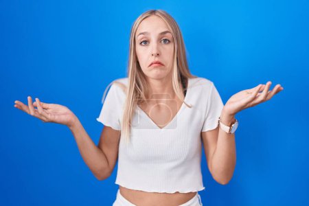 Photo for Young caucasian woman standing over blue background clueless and confused expression with arms and hands raised. doubt concept. - Royalty Free Image
