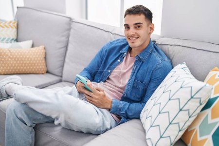 Photo for Young hispanic man using smartphone sitting on sofa at home - Royalty Free Image