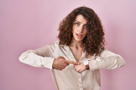 Téléchargez les photos : Hispanic woman with curly hair standing over pink background in hurry pointing to watch time, impatience, upset and angry for deadline delay - en image libre de droit