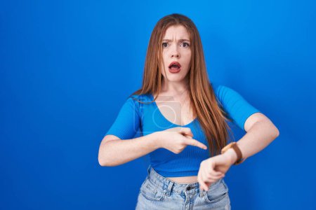 Téléchargez les photos : Redhead woman standing over blue background in hurry pointing to watch time, impatience, upset and angry for deadline delay - en image libre de droit