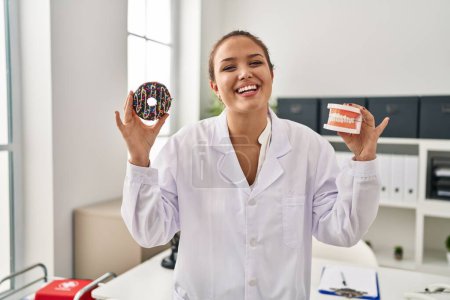 Photo for Young hispanic dentist woman holding denture and chocolate doughnut smiling and laughing hard out loud because funny crazy joke. - Royalty Free Image