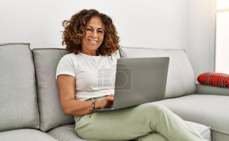 Téléchargez les photos : Moyen Age femme hispanique souriant confiant en utilisant un ordinateur portable à la maison - en image libre de droit