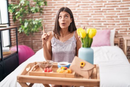 Téléchargez les photos : Brunette young woman eating breakfast sitting on the bed amazed and surprised looking up and pointing with fingers and raised arms. - en image libre de droit