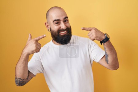 Téléchargez les photos : Jeune homme hispanique avec barbe et tatouages debout sur fond jaune souriant gai montrant et pointant avec les doigts et la bouche. concept de santé dentaire. - en image libre de droit