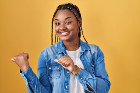 Foto de African american woman with braids standing over yellow background pointing to the back behind with hand and thumbs up, smiling confident - Imagen libre de derechos