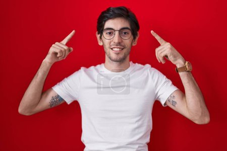 Foto de Joven hombre hispano de pie sobre fondo rojo sonriendo señalando a la cabeza con ambas manos dedo, gran idea o pensamiento, buena memoria - Imagen libre de derechos