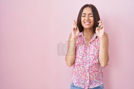 Téléchargez les photos : Young hispanic woman with long hair standing over pink background gesturing finger crossed smiling with hope and eyes closed. luck and superstitious concept. - en image libre de droit