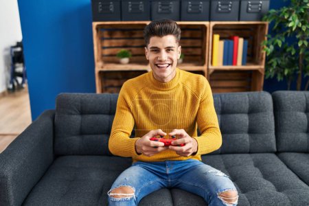 Foto de Young hispanic man playing video game sitting on sofa at home - Imagen libre de derechos