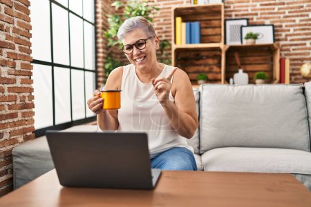 Photo for Middle age woman using laptop at home doing video call smiling happy pointing with hand and finger to the side - Royalty Free Image