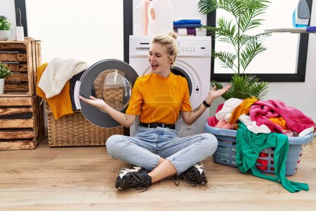 Téléchargez les photos : Young blonde woman doing laundry sitting by washing machine smiling showing both hands open palms, presenting and advertising comparison and balance - en image libre de droit