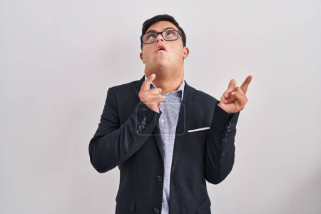 Photo for Young hispanic man with down syndrome wearing business style amazed and surprised looking up and pointing with fingers and raised arms. - Royalty Free Image