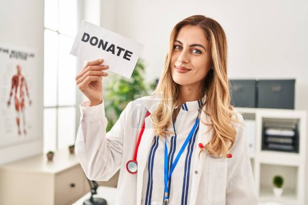 Photo for Young blonde doctor woman supporting organs donations looking positive and happy standing and smiling with a confident smile showing teeth - Royalty Free Image
