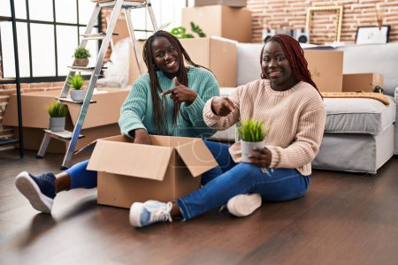 Téléchargez les photos : Two african woman moving to a new home sitting on the floor smiling happy pointing with hand and finger - en image libre de droit