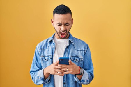 Photo for Young hispanic man using smartphone typing message angry and mad screaming frustrated and furious, shouting with anger. rage and aggressive concept. - Royalty Free Image