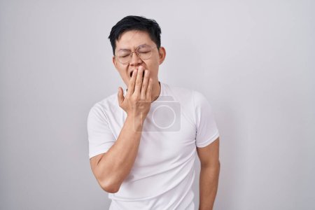 Foto de Young asian man standing over white background bored yawning tired covering mouth with hand. restless and sleepiness. - Imagen libre de derechos