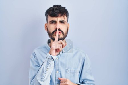 Foto de Young hispanic man with beard standing over blue background asking to be quiet with finger on lips. silence and secret concept. - Imagen libre de derechos