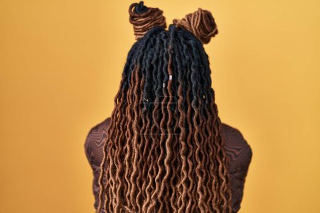 Foto de African woman with braided hair standing over yellow background standing backwards looking away with crossed arms - Imagen libre de derechos