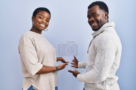 Foto de Joven pareja afroamericana de pie sobre fondo azul juntos invitando a entrar sonriente natural con la mano abierta - Imagen libre de derechos