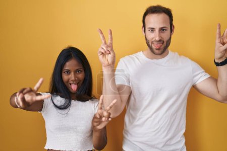 Téléchargez les photos : Couple Interracial debout sur fond jaune souriant avec la langue montrant les doigts des deux mains faisant signe de victoire. numéro deux. - en image libre de droit